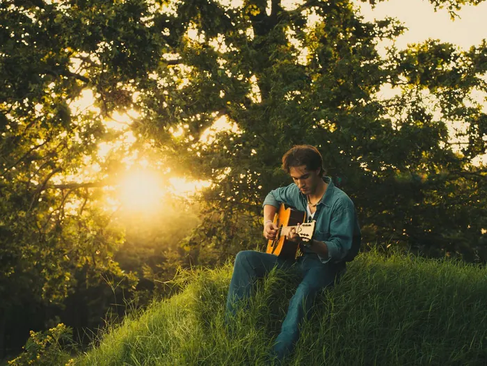 Senior playing guitar on a grassy hill with the sun behind him
