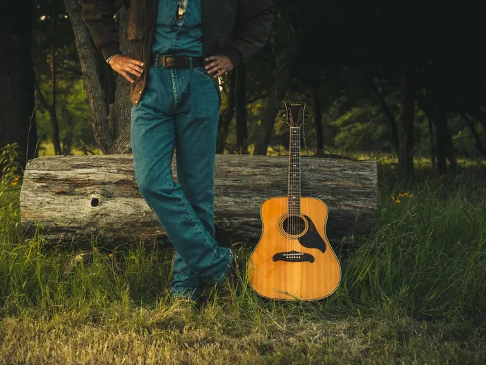 Senior in jeans leaning on a log beside his guitar in the woods